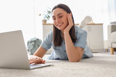 Genç bir kadın evdeki yumuşak halıda dizüstü bilgisayar kullanıyor.