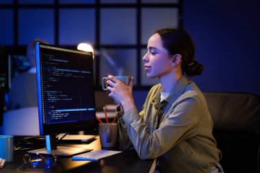 Elinde kahve fincanı olan kadın programcı gece ofiste oturuyor.