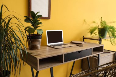 Ofiste sarı duvarın yanında dizüstü bilgisayarı ve bitkileri olan modern bir iş yeri.