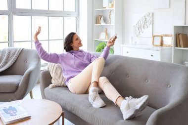 Oturma odasındaki kanepede uzanmış, uzaktan kumandalı, gülümseyen genç bir kadın.