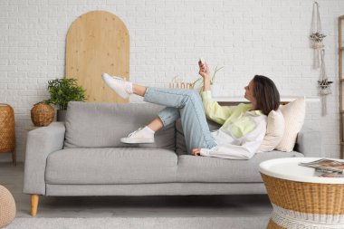 Oturma odasındaki kanepede uzanmış, uzaktan kumandalı, gülümseyen genç bir kadın.