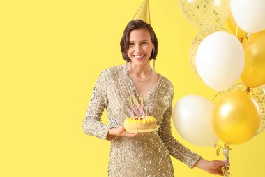 Doğum günü pastası ve sarı arka planda balonları olan parti şapkalı güzel kadın.