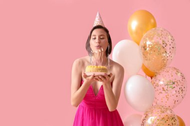Doğum günü pastası ve balonlarıyla parti şapkalı güzel bir kadın pembe arka planda dilek diliyor