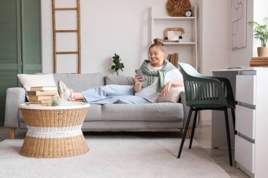 Gözlüklü genç kadın kanepede uzanıyor ve oturma odasında cep telefonuyla sohbet ediyor.