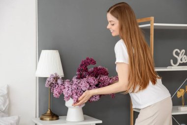 Evdeki komodinin üzerinde leylak demeti olan genç bir kadın.