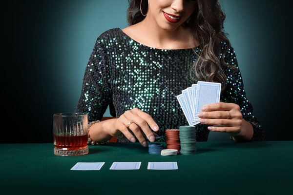 Beautiful young African-American woman sitting at poker table with playing cards, chips and glass of whiskey on dark green background