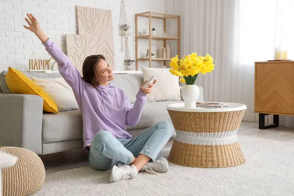 Smiling young woman with air conditioner remote control sitting near sofa in living room