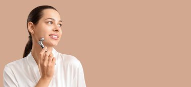 Beautiful young woman massaging her face with roller on beige background with space for text