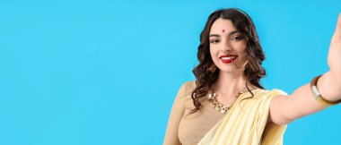 Beautiful young Indian woman in sari taking selfie on blue background with space for text