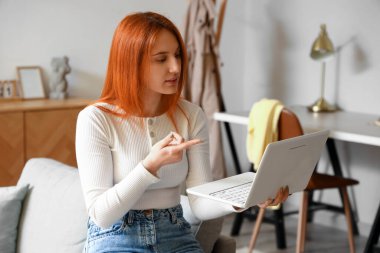 Dizüstü bilgisayarlı genç bir kadın evdeki kanepede sohbet ediyor.