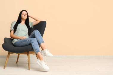 Güzel genç bayan bej duvarın yanındaki rahat koltukta dinleniyor.