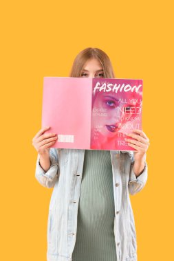 Beautiful young pregnant woman with magazine on yellow background