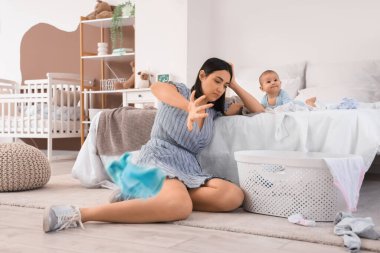 Young woman with laundry and her baby suffering from postnatal depression in bedroom