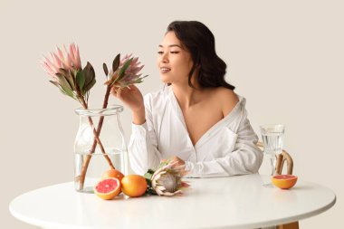 Beyaz duvarın yanındaki masada güzel protea çiçekleri ve taze greyfurtlu genç Asyalı kadın.