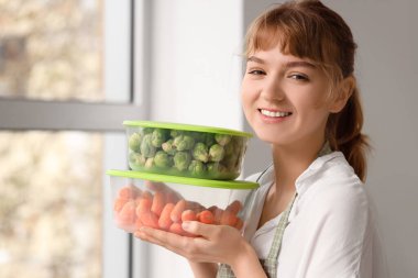 Beautiful young woman holding plastic containers with frozen vegetables in kitchen, closeup