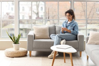 Genç Afro-Amerikalı kadın evdeki koltukta cep telefonu kullanıyor.