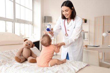 Female pediatrician measuring temperature of little baby on couch in clinic