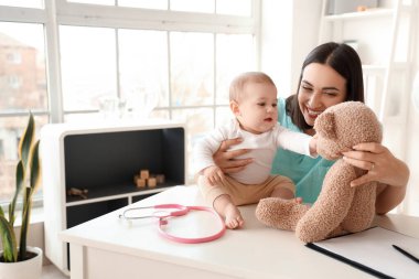 Female pediatrician with little baby and toy bear at table in clinic