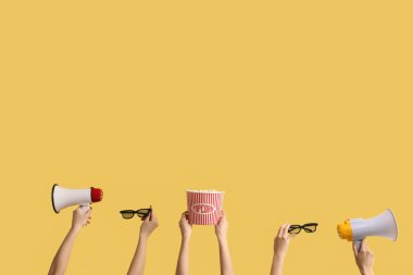 Hands holding bucket with popcorn, 3D glasses and megaphones on yellow background
