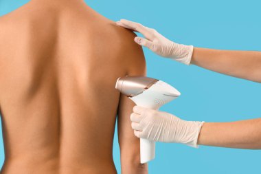 Kozmetik uzmanı mavi arka planda insan vücudundan saç çıkarmak için fotoepilatör kullanıyor.