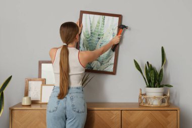 Young woman hanging painting on light wall at home, back view