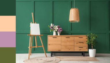 Stylish wooden cabinet, easel and vase with flowers near green wall in room. Different color patterns