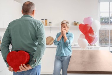 Mature man hiding gift for his wife behind back in kitchen on Valentine's Day