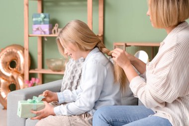 Beautiful young woman doing hair her cute little daughter with gift box and sitting on sofa in living room