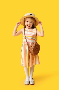 Cute little girl in hat with stylish wicker bag on yellow background