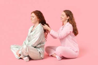 Happy redhead woman brushing her sister's hair on pink background