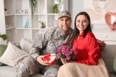 Man in military uniform greeting his wife with flowers and heart-shaped gift box at home. Valentine's Day celebration