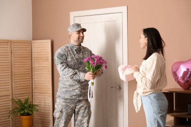 Man in military uniform greeting his wife with flowers and heart-shaped gift box at home. Valentine's Day celebration