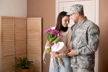 Man in military uniform greeting his wife with flowers and heart-shaped gift box at home. Valentine's Day celebration
