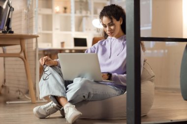 Female African-American programmer with eyeglasses and laptop on beanbag in office at night