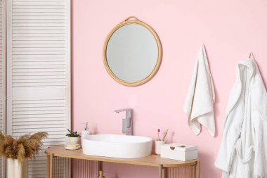 Modern sink with bath supplies and houseplant on shelving unit near pink wall in bathroom