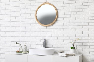 Banyodaki beyaz tuğla duvarın yanındaki tezgahta banyo malzemeleri ve güzel çiçeklerle modern lavabo.