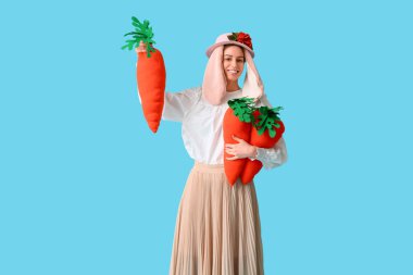 Beautiful young woman in bunny ears holding toy carrots on blue background. Easter celebration