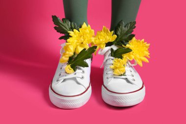 Pembe arka planda sarı kasımpatı çiçekleri olan beyaz ayakkabılı güzel genç kadın bacakları.