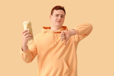 Young man holding tasty doner kebab and showing thumb-down on beige background