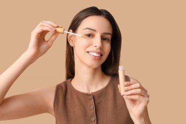 Beautiful young woman applying concealer on brown background