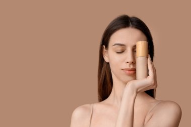 Beautiful young woman covering one eye by foundation on brown background