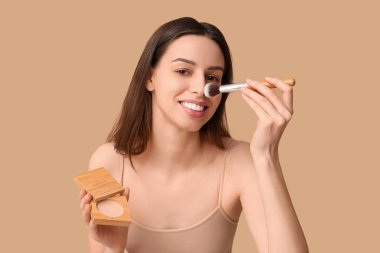 Beautiful young woman with brush applying powder on brown background