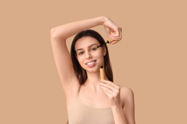 Beautiful young woman applying mascara on brown background