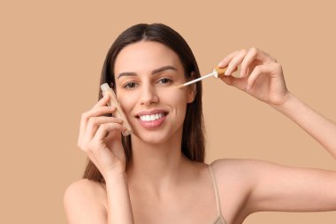 Beautiful young woman applying concealer on brown background
