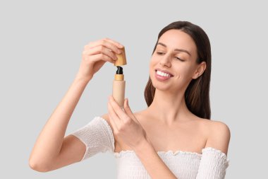 Beautiful young woman opening foundation on grey background