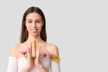 Beautiful young woman with flowers holding foundation on grey background