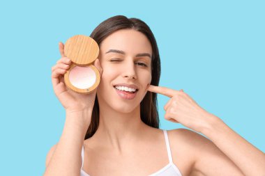 Beautiful young woman with highlighter pointing at cheek on blue background