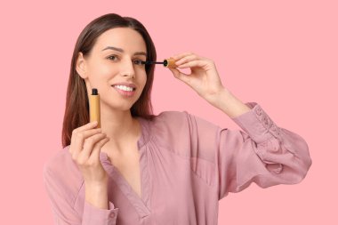Beautiful young woman applying mascara on pink background