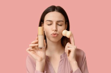 Beautiful young woman holding makeup sponge and face foundation on pink background