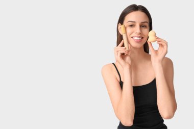 Beautiful young woman holding makeup sponge and concealer on white background